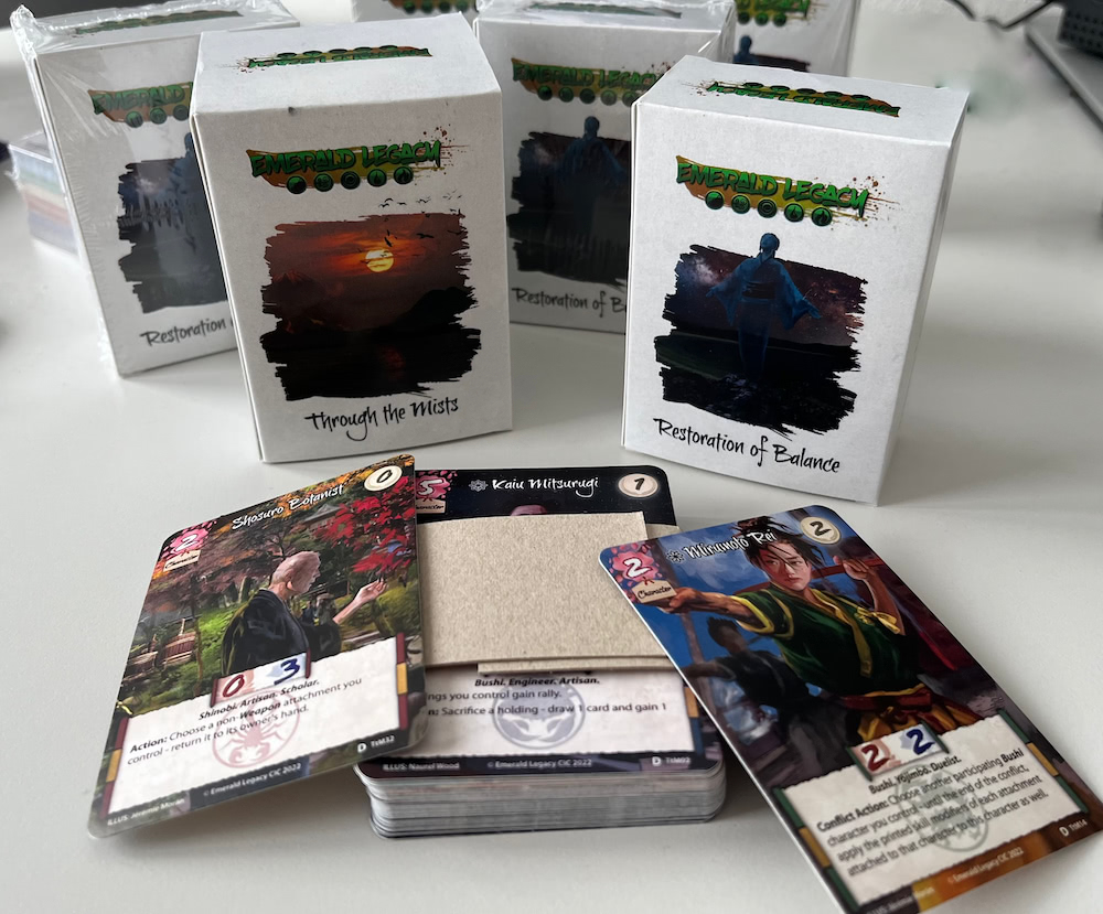 Photo of several tuck boxes of Restoration of Balance and Through the Mists, with three cards of the Game Night Kit laying in front of the boxes.